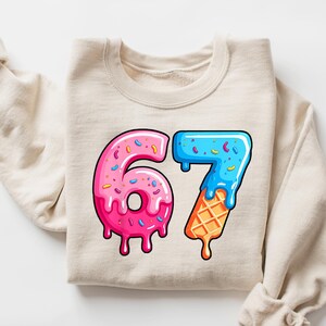 May include: Beige sweatshirt featuring the number 67 in a colourful design. The number 6 is a pink donut with sprinkles, and the number 7 is a blue ice cream cone with a waffle cone base. The design has a dripping effect, perfect for a birthday.