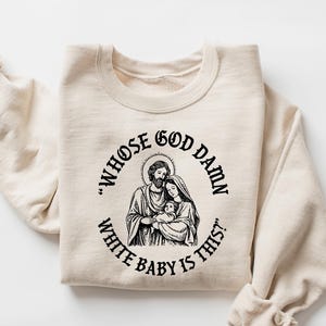 May include: Cream-colored sweatshirt with a black graphic of a religious family and the text "WHOSE GOD DAMN WHITE BABY IS THIS?" The sweatshirt is made of a soft material and has long sleeves.
