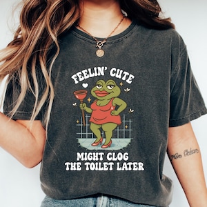 Puede incluir: Camiseta gris oscuro con un gráfico de una rana con un vestido rojo sosteniendo un desatascador. El texto en la camiseta dice "FEELIN' CUTE MIGHT CLOG THE TOILET LATER". La camiseta se usa con vaqueros azules y zapatillas blancas.