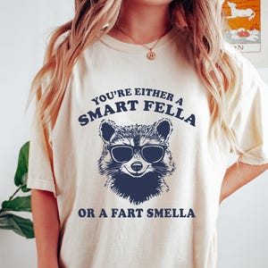 May include: Cream-colored t-shirt with a raccoon wearing sunglasses graphic. The text reads "You're either a Smart Fella or a Fart Smella." The shirt is part of a handmade gifts collection.