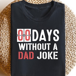 May include: Black t-shirt with the text "00 DAYS WITHOUT A DAD JOKE" in white and red lettering. The shirt is displayed on a round, woven surface. The text is in a bold, sans-serif font.