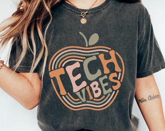 May include: Dark gray t-shirt with a colorful apple design. The apple graphic features the words "TECH VIBES" in orange, pink, and green. The shirt is paired with blue jeans and white sneakers.