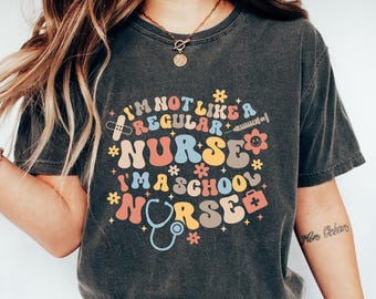 May include: Dark gray t-shirt with the text "I'M NOT LIKE A REGULAR NURSE I'M A SCHOOL NURSE" in a colorful, retro design. The design includes medical symbols like a stethoscope, syringe, and bandage. The shirt is paired with blue jeans and white sneakers.
