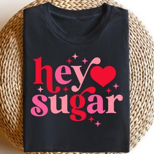 Puede incluir: Camiseta negra con un gráfico rosa y rojo que dice "hey sugar" con un corazón en el medio.