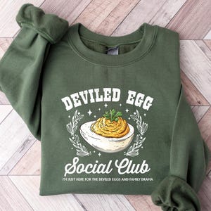 May include: A dark green sweatshirt with the words "Deviled Egg Social Club" in white, along with an illustration of a deviled egg. The text "I'm just here for the deviled eggs and family drama" is printed below the graphic.