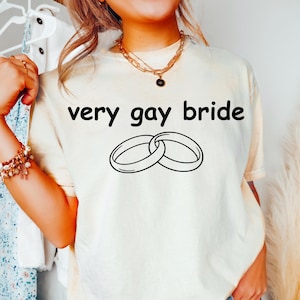 Puede incluir: Camiseta color crema con la frase "very gay bride" y dos anillos entrelazados. El texto y los anillos son negros. La camiseta tiene un corte relajado y un color neutro, ideal para una boda o celebración nupcial.