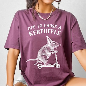 Peut inclure: T-shirt mauve avec l'inscription "OFF TO CAUSE A KERFUFFLE" et un dessin blanc d'un rat sur un scooter avec un chapeau de fête. Le t-shirt est à manches courtes.