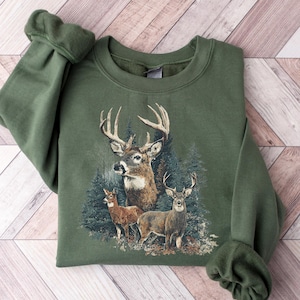 Könnte beinhalten: Ein olivgrünes Sweatshirt mit einer detaillierten Grafik von Hirschen in einem Wald. Das Design umfasst einen Hirsch mit großem Geweih und zwei kleinere Hirsche. Das Sweatshirt hat einen Rundhalsausschnitt und lange Ärmel mit gerippten Bündchen.