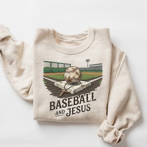 Puede incluir: Una sudadera de cuello redondo color crema con una ilustración gráfica de una pelota de béisbol sobre una base de estadio. Una pequeña cruz de madera cuelga de la pelota. El texto "BASEBALL AND JESUS" está impreso debajo del gráfico.