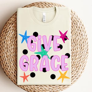 May include: A cream-coloured t-shirt featuring the text "GIVE GRACE" in large, light pink bubble letters. The design includes a pattern of black polka dots and several hand-drawn style stars in bright blue, hot pink, teal, orange, and red.