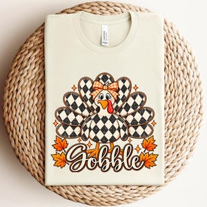 May include: A cream-colored t-shirt featuring a graphic of a turkey with a black and white checkered pattern on its body and tail feathers. The turkey wears an orange and white checkered bow. The word Gobble is printed below in brown.