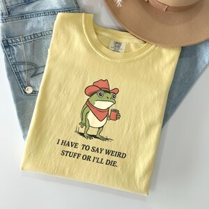 Puede incluir: Camiseta amarilla clara con un dibujo de una rana con sombrero de vaquero y una taza. El texto "I HAVE TO SAY WEIRD STUFF OR I'LL DIE" está impreso debajo. La camiseta se combina con vaqueros azul claro y un sombrero beige.