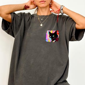 Può includere: T-shirt grigio scuro oversize con una tasca con una grafica colorata di un gatto nero. Il gatto ha occhi blu brillante ed è ambientato su uno sfondo astratto e vibrante. La maglietta ha maniche corte e una vestibilità ampia.