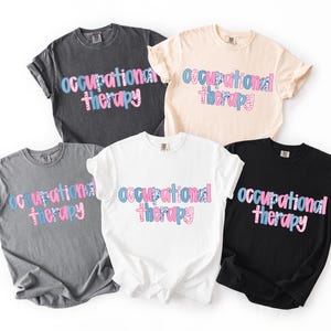 Könnte beinhalten: Fünf kurzärmelige T-Shirts mit Rundhalsausschnitt in Schwarz, Weiß, Grau, Dunkelgrau und Creme. Jedes Shirt zeigt den Text "occupational therapy" in einer stilisierten Schriftart mit einem blau-rosa Blumenmuster auf den Buchstaben, angeordnet in zwei Zeilen.