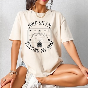 Könnte beinhalten: Ein beiges Kurzarm-T-Shirt mit einem schwarzen Grafikdruck eines Spirit-Boards, dem Text "HOLD ON I'M TEXTING MY MOM" in einem Bogen darüber und darunter sowie einem Planchette-Symbol unterhalb des Alphabets und der Zahlen.