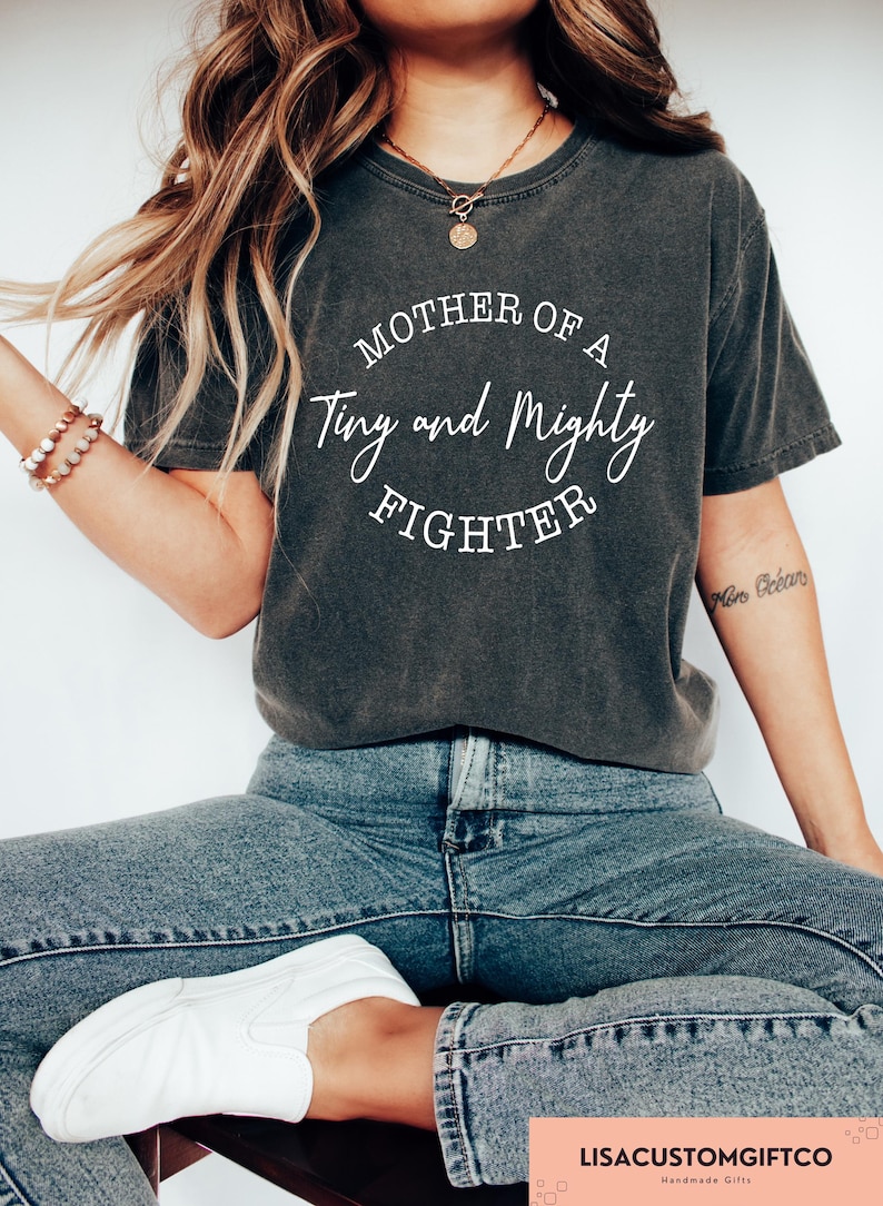 May include: A dark grey t-shirt with the text "Mother of a Tiny and Mighty Fighter" in white lettering.