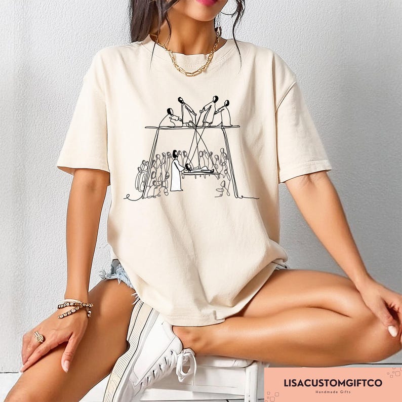 May include: Beige short-sleeved t-shirt featuring a black line drawing of a religious scene. The graphic depicts figures on a platform, with others below. The text "LISACUSTOMGIFTCO Handmade Gifts" is visible at the bottom right.