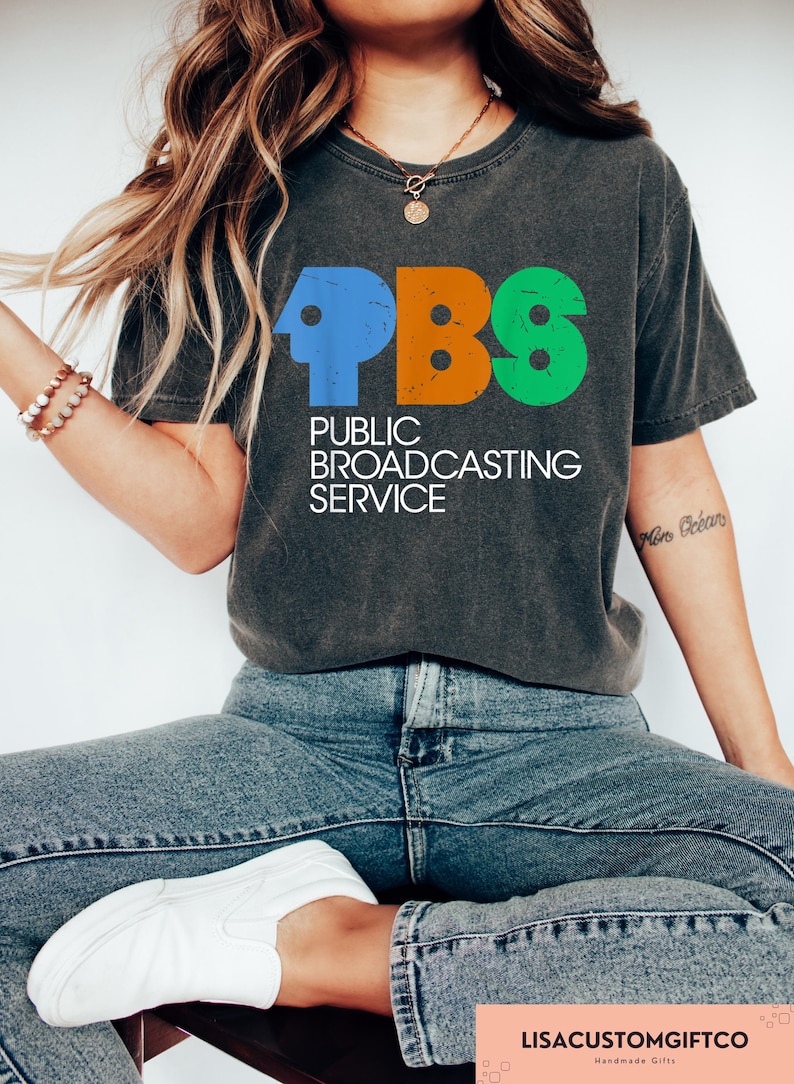 May include: Dark grey t-shirt with the PBS logo in blue, orange, and green, and the words "PUBLIC BROADCASTING SERVICE" in white. The shirt is a casual style.