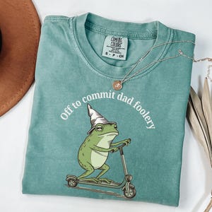 Op de afbeelding: Een turquoise Comfort Colors T-shirt met een cartoon kikker op een scooter, met een tovenaars hoed. De tekst "Off to commit dad foolery" is boven de kikker gedrukt. Een zilveren ketting met een kleine hanger ligt over het T-shirt.