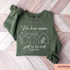 May include: Olive green sweatshirt with white graphic text and illustrations. The text reads "He has risen just as he said. Luke 24:6." The graphic includes a cross, a bow, and an empty tomb. The sweatshirt is folded on a light wood surface.