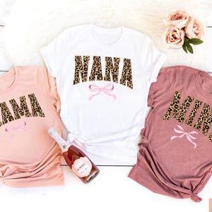 Peut inclure: Trois t-shirts avec un motif léopard et les mots "Mama", "Nana" et "Mini" en lettres roses avec un nœud rose sous chaque mot.