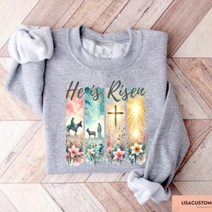 May include: A gray sweatshirt with a colorful graphic print of a cross, flowers, and a scene from the Bible. The text "He is Risen" is printed in black on the sweatshirt.