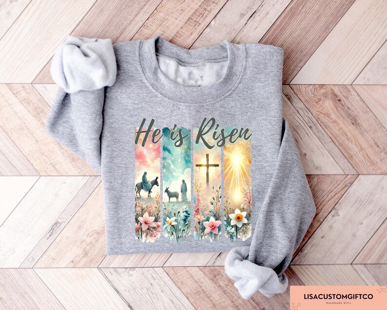 May include: A gray sweatshirt with a colorful graphic print of a cross, flowers, and a scene from the Bible. The text "He is Risen" is printed in black on the sweatshirt.