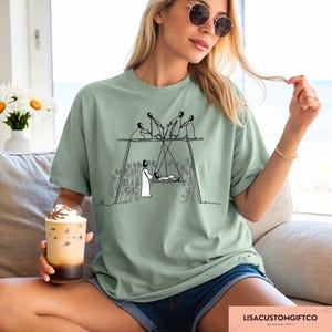 May include: Sage green t-shirt featuring a black and white line drawing of a religious scene. The graphic depicts figures on a structure above a person lying down, with a crowd below. The text "LISACUSTOMGIFTCO" is at the bottom.