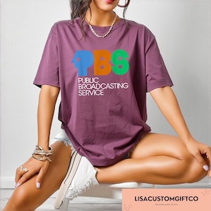 May include: A maroon t-shirt with the PBS logo in blue, orange, and green, and the words "PUBLIC BROADCASTING SERVICE" in white. The shirt is paired with white shorts and trainers.