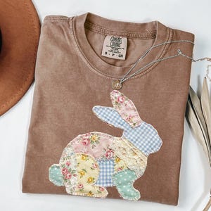 以下が含まれることがあります： パッチワークのウサギのデザインが施された茶色のComfort ColorsのTシャツ。ウサギは、パステルカラーの花柄、ギンガムチェック、水玉模様が特徴です。小さなペンダントが付いたシルバーのネックレスがシャツの上にあります。シャツは白い面に折りたたまれています。