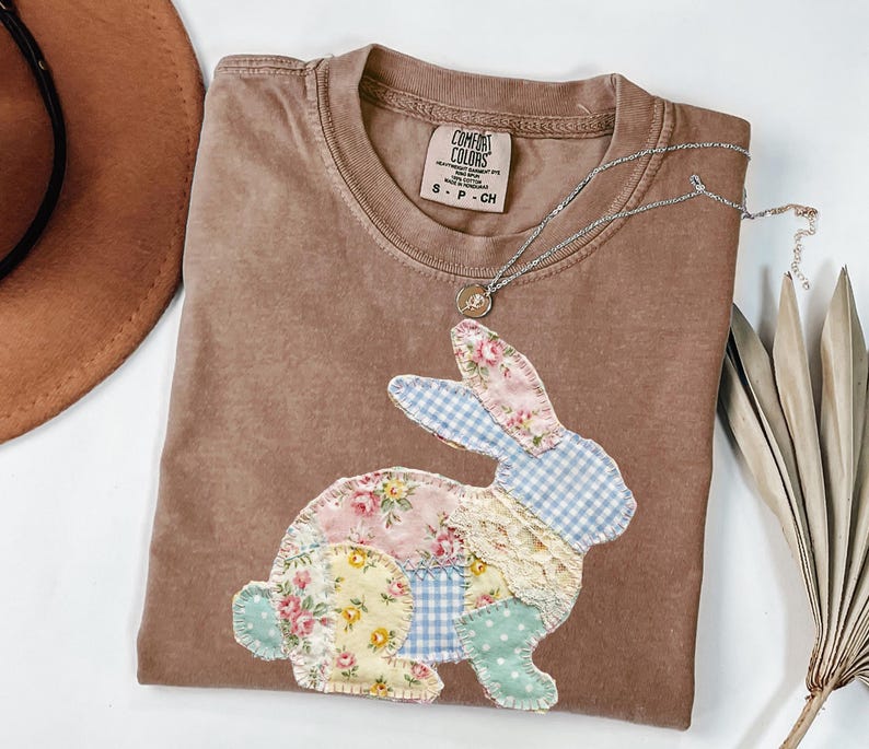 以下が含まれることがあります： パッチワークのウサギのデザインが施された茶色のComfort ColorsのTシャツ。ウサギは、パステルカラーの花柄、ギンガムチェック、水玉模様が特徴です。小さなペンダントが付いたシルバーのネックレスがシャツの上にあります。シャツは白い面に折りたたまれています。
