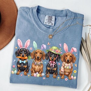 May include: A blue Comfort Colors t-shirt featuring an Easter-themed graphic of four dachshunds wearing bunny ears and holding Easter eggs. The dogs are adorned with accessories like bow ties and hats. A gold necklace is draped across the shirt.