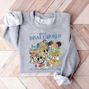 May include: A gray sweatshirt featuring a colorful graphic of Disney characters and attractions. The text "Walt Disney World" is displayed above the graphic, with "The Most Magical Place On Earth" below. The sweatshirt has rolled-up sleeves.