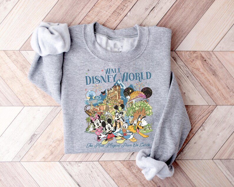 May include: A gray sweatshirt featuring a colorful graphic of Disney characters and attractions. The text "Walt Disney World" is displayed above the graphic, with "The Most Magical Place On Earth" below. The sweatshirt has rolled-up sleeves.