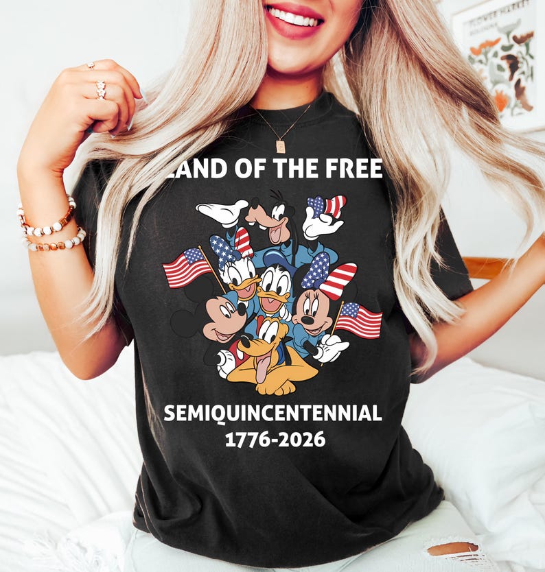 May include: A black short-sleeve t-shirt featuring a graphic of Disney characters including Mickey Mouse, Minnie Mouse, Donald Duck, Daisy Duck, Goofy, and Pluto holding American flags. Text reads LAND OF THE FREE SEMIQUINCENTENNIAL 1776-2026.