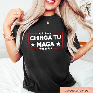 May include: Black t-shirt featuring the text "CHINGA TU MAGA" in white, within a red rectangle and white stars. The shirt has a crew neck design.