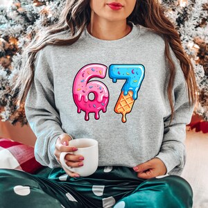 May include: A grey sweatshirt featuring the number 67 in a colourful design. The number 6 is a pink donut with sprinkles, and the 7 is a blue ice cream cone. The person is holding a white mug.