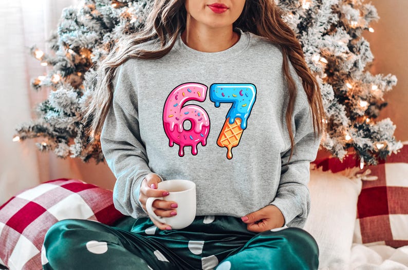 May include: A grey sweatshirt featuring the number 67 in a colourful design. The number 6 is a pink donut with sprinkles, and the 7 is a blue ice cream cone. The person is holding a white mug.