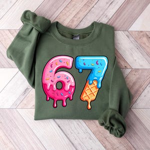 May include: An olive green sweatshirt with a vibrant graphic of the number 67. The number 6 is a pink donut with sprinkles, and the 7 is a blue ice cream cone with a waffle cone base. The design has a dripping effect, creating a playful aesthetic.