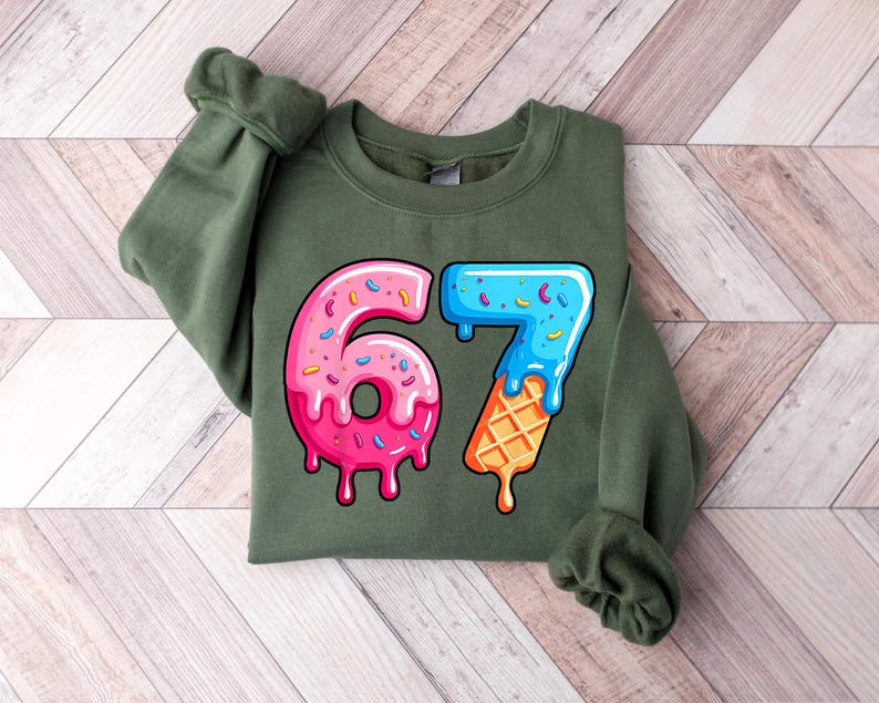 May include: An olive green sweatshirt with a vibrant graphic of the number 67. The number 6 is a pink donut with sprinkles, and the 7 is a blue ice cream cone with a waffle cone base. The design has a dripping effect, creating a playful aesthetic.