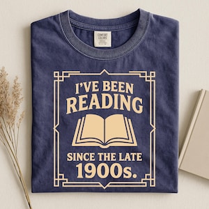 May include: A navy blue crew neck t-shirt featuring a cream-colored graphic print. The design includes a rectangular border, an open book icon, and the text: I'VE BEEN READING SINCE THE LATE 1900s. The shirt has a Comfort Colors brand neck label.
