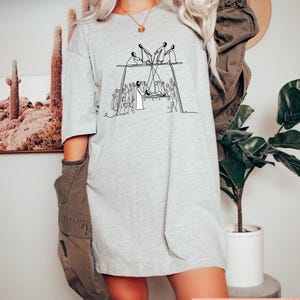May include: A light gray t-shirt with a black line drawing of a group of people on a swing set. The shirt has short sleeves and a relaxed fit. The text "LISACUSTOMGIFTCO" is printed in a pink rectangle at the bottom right.