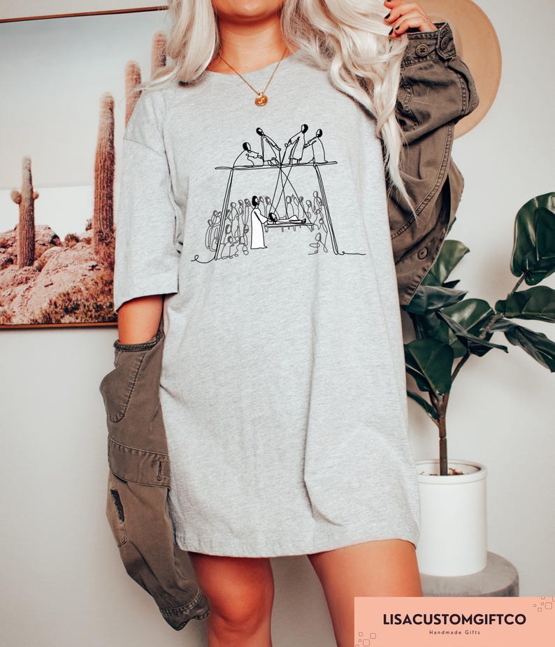 May include: A light gray t-shirt with a black line drawing of a group of people on a swing set. The shirt has short sleeves and a relaxed fit. The text "LISACUSTOMGIFTCO" is printed in a pink rectangle at the bottom right.