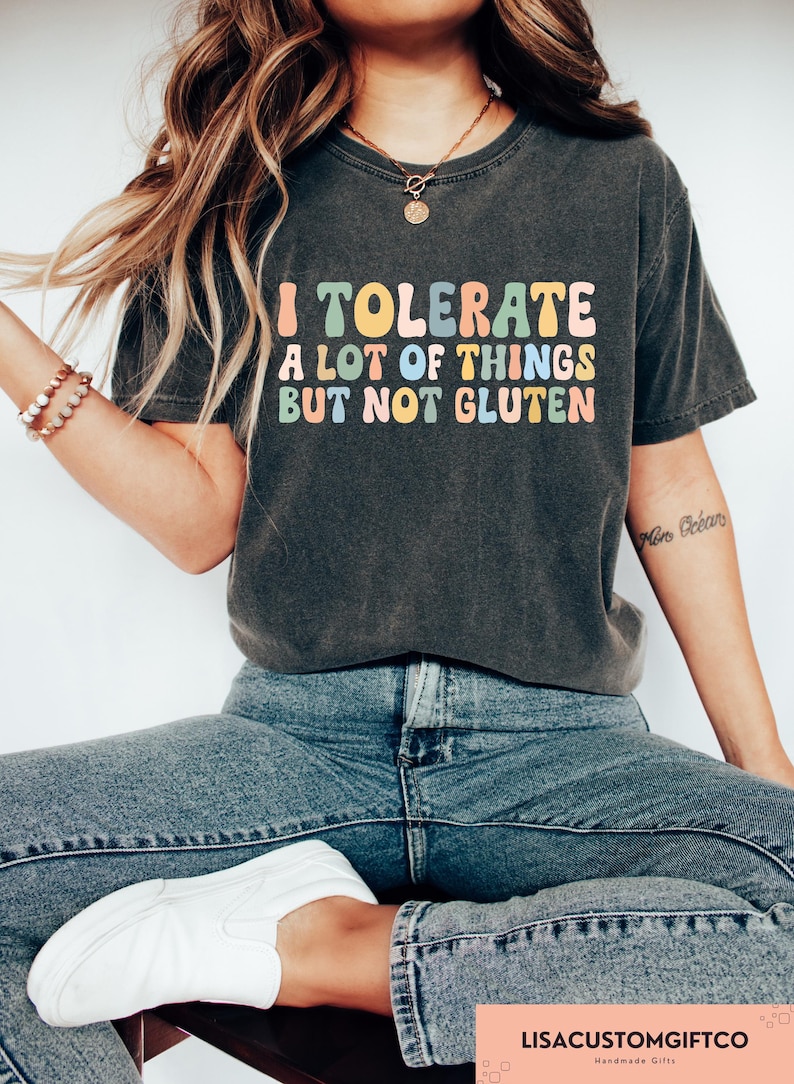 K&ouml;nnte beinhalten: Ein graues T-Shirt mit dem Text "I tolerate a lot of things but not gluten" in bunten Buchstaben. Das Shirt wird von einer Person getragen, die blaue Jeans und wei&szlig;e Turnschuhe tr&auml;gt.