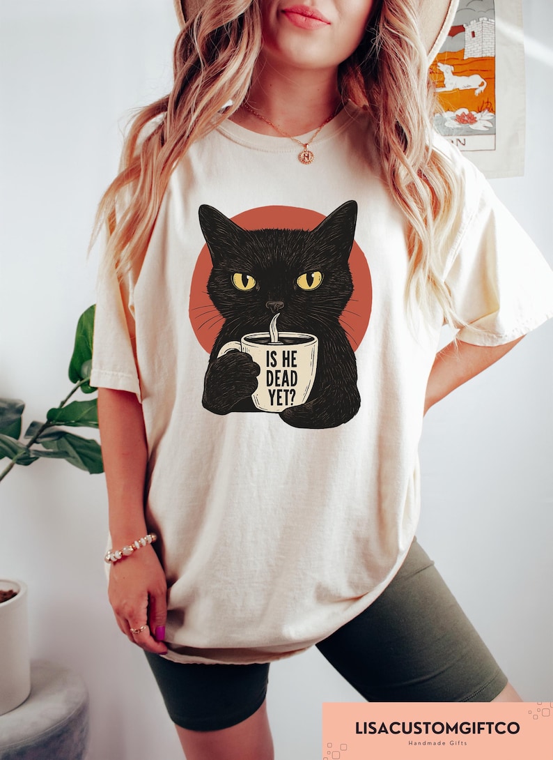 May include: Cream-colored t-shirt featuring a black cat illustration with yellow eyes, holding a coffee mug. The mug has the text "IS HE DEAD YET?". The cat is set against a red circle.