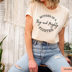 May include: A woman wearing a light beige t-shirt with the text "Mother of a Tiny and Mighty Fighter" printed on it. She is wearing blue jeans and is standing in front of a white wooden wall.