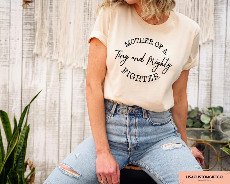 May include: A woman wearing a light beige t-shirt with the text "Mother of a Tiny and Mighty Fighter" printed on it. She is wearing blue jeans and is standing in front of a white wooden wall.