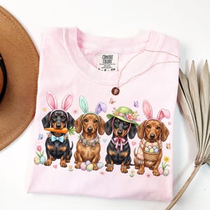 May include: A light pink Comfort Colors t-shirt featuring four dachshunds dressed for Easter. Each dog wears bunny ears or a hat, and is surrounded by Easter eggs, flowers, and butterflies. The shirt also has a Comfort Colors tag.