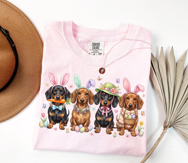 May include: A light pink Comfort Colors t-shirt featuring four dachshunds dressed for Easter. Each dog wears bunny ears or a hat, and is surrounded by Easter eggs, flowers, and butterflies. The shirt also has a Comfort Colors tag.