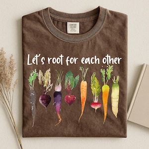 Puede incluir: Una camiseta marrón con cuello redondo y una etiqueta Comfort Colors. El frente presenta un gráfico de ocho coloridas hortalizas de raíz, incluyendo zanahorias y remolachas, con el texto blanco "Let's root for each other" impreso encima.